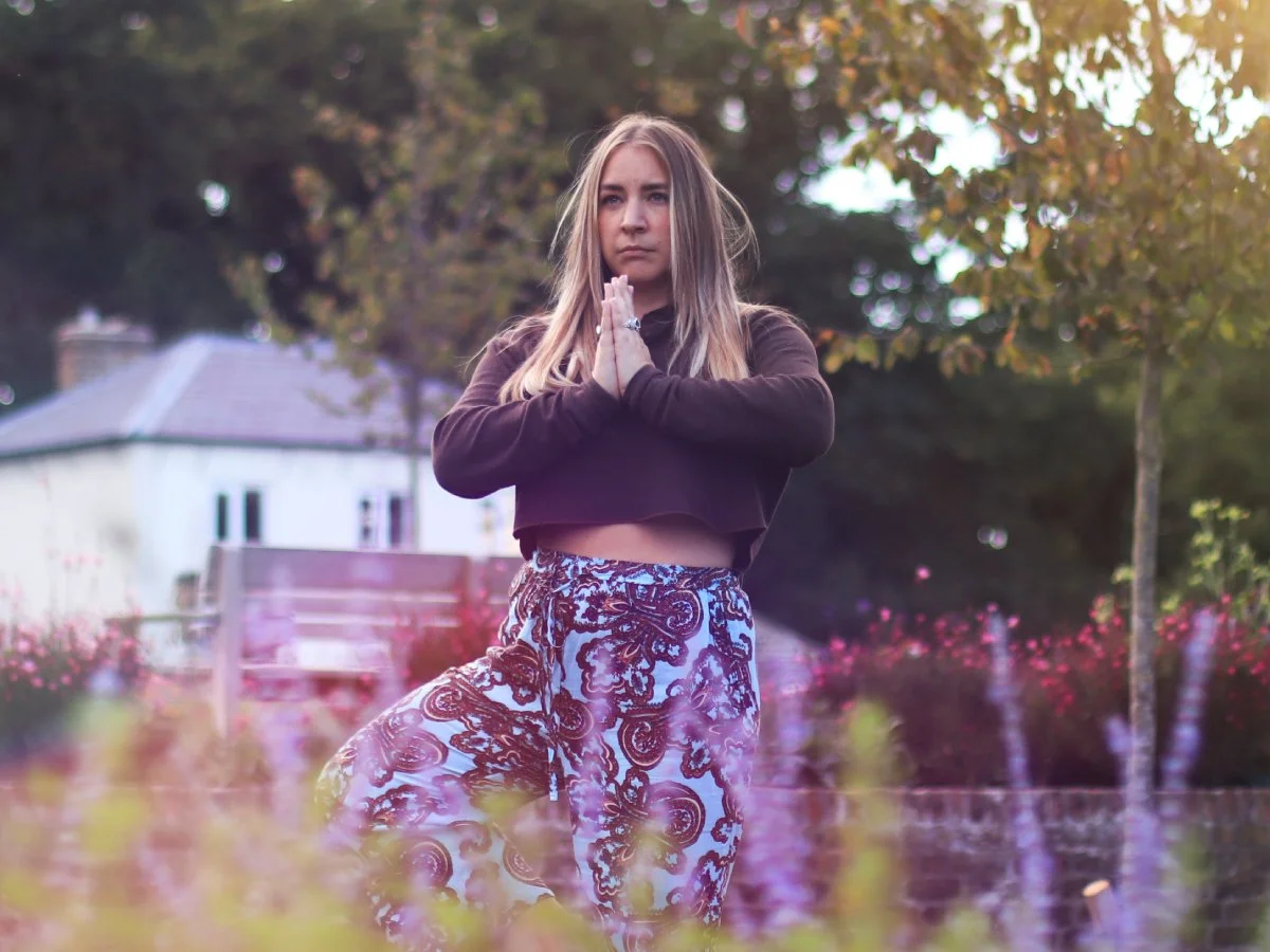 A young woman practicing yoga outdoors in a garden during sunset, standing with hands in prayer position, wearing a black long-sleeved top and patterned pants.