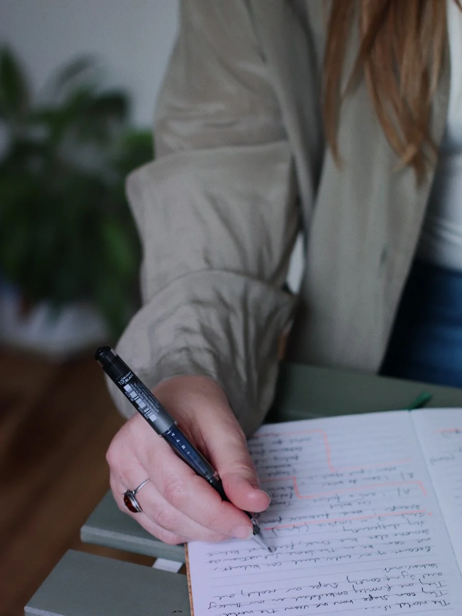 A person writing with a black pen in a notebook with handwritten notes, dressed in a beige jacket, with a plant in the background.