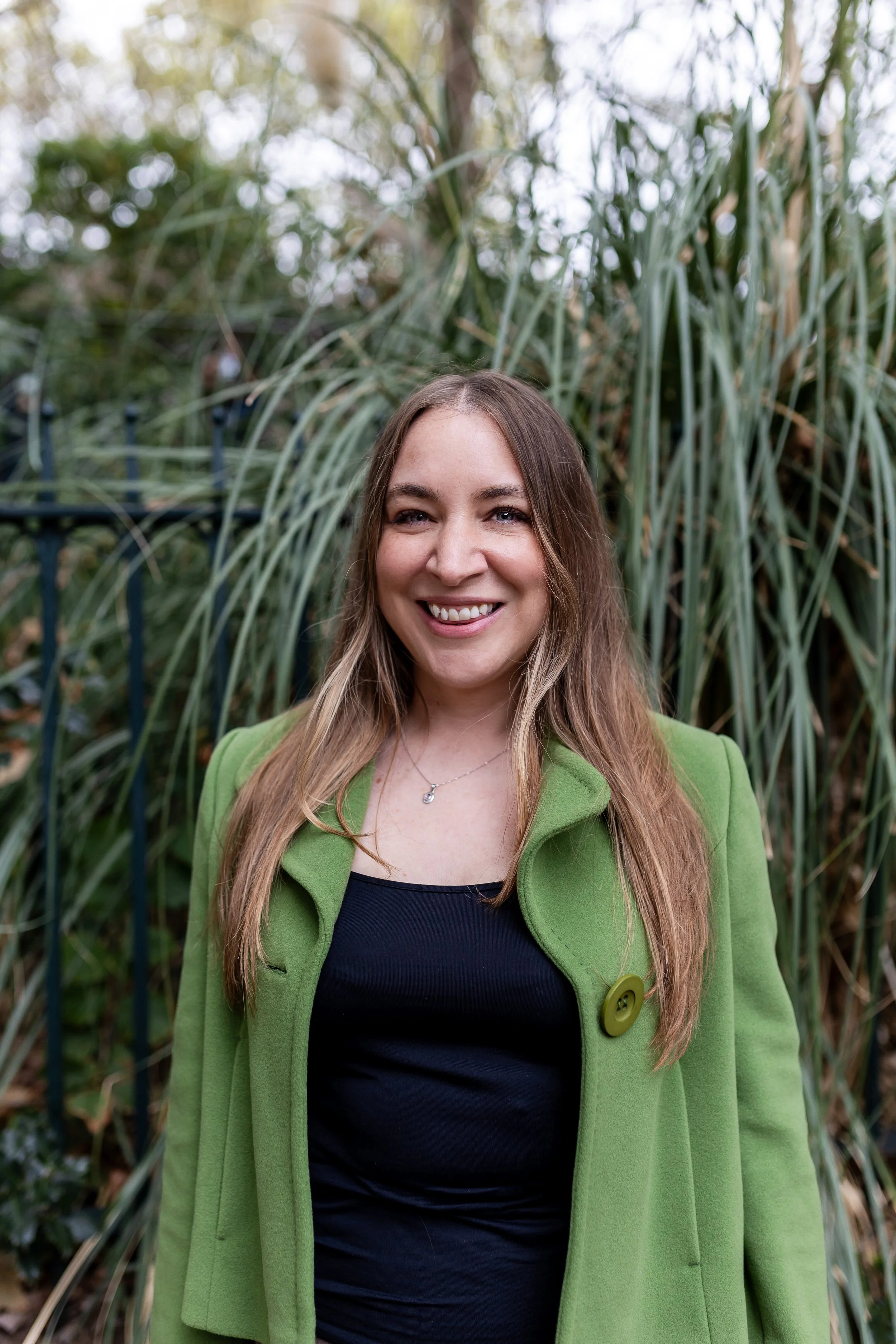 Portrait of a woman smiling outdoors with tall grass and trees in the background, wearing a green coat over a black top.