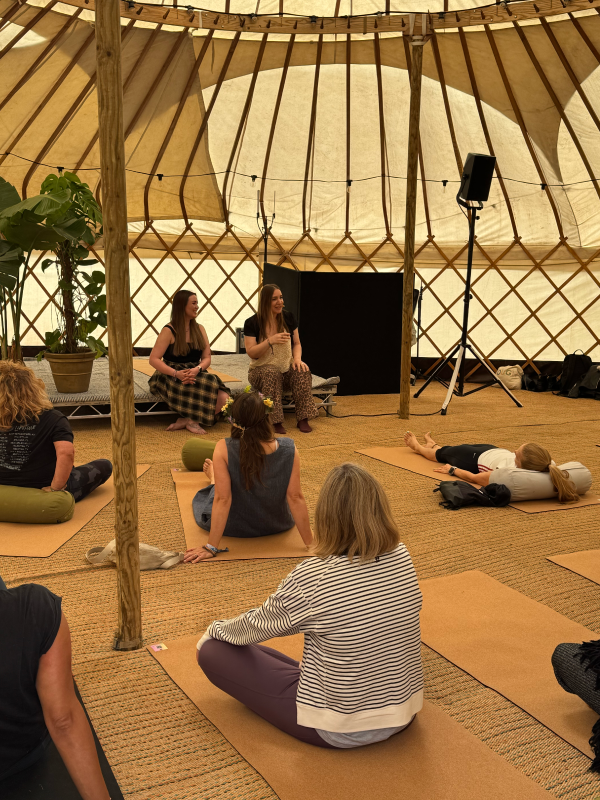 People participating in a yoga or meditation session inside a large, beige fabric yurt, with two women speaking at the front and participants sitting or lying on yoga mats.