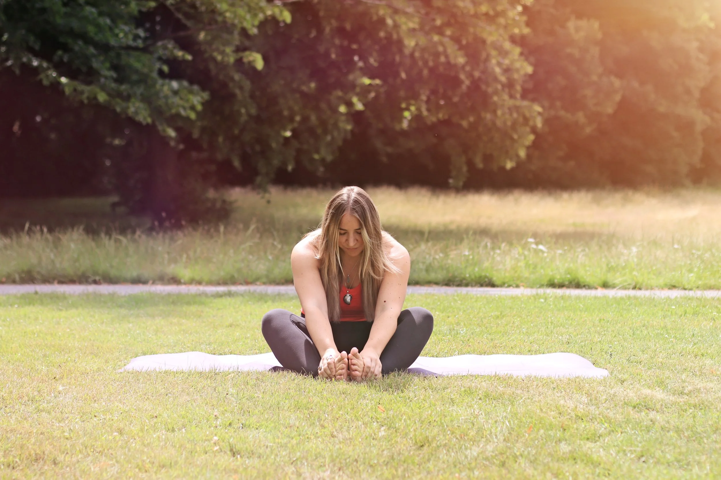 A woman practicing yoga outdoors on a white mat in a park, sitting in a forward bend stretch with legs crossed and leaning forward to touch her toes, during daytime with sunlight filtering through trees.