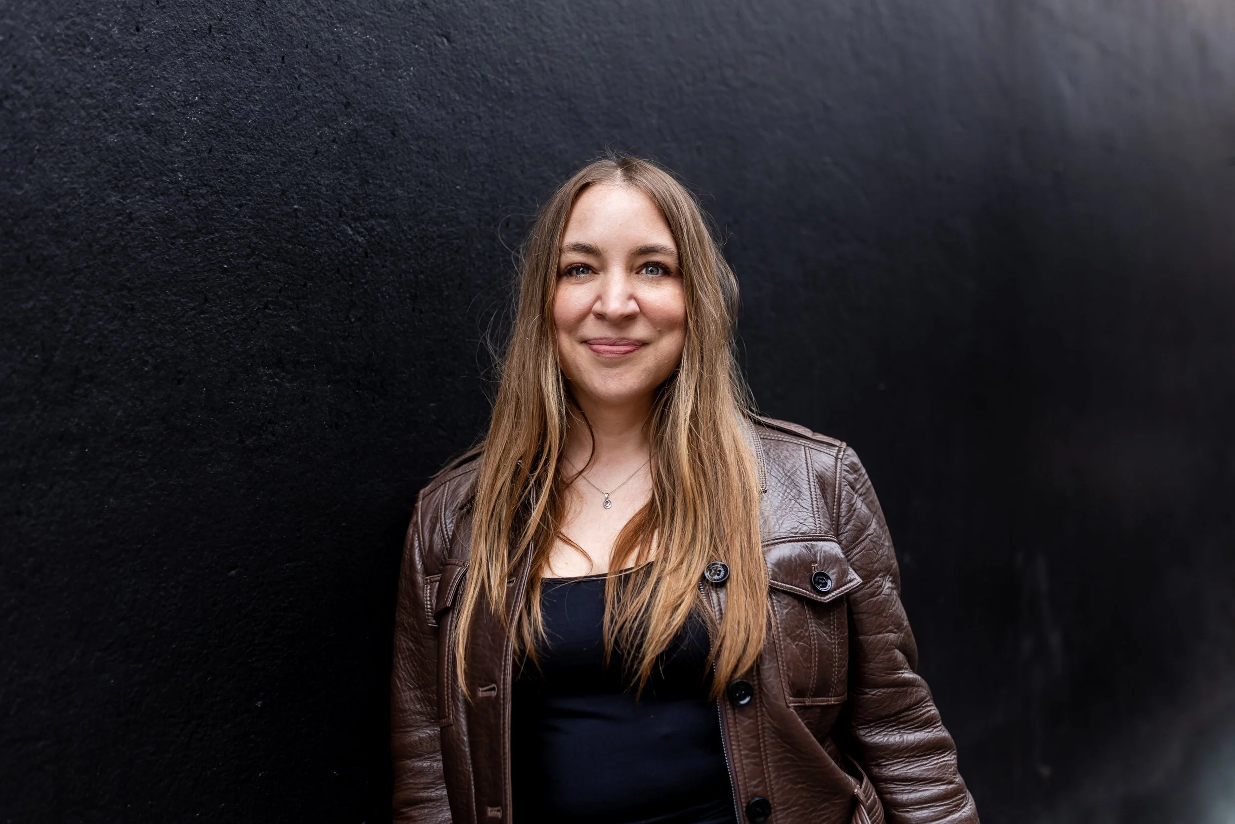 A woman with long, wavy, light brown hair smiling and standing in front of a textured black wall, wearing a brown leather jacket over a black top.