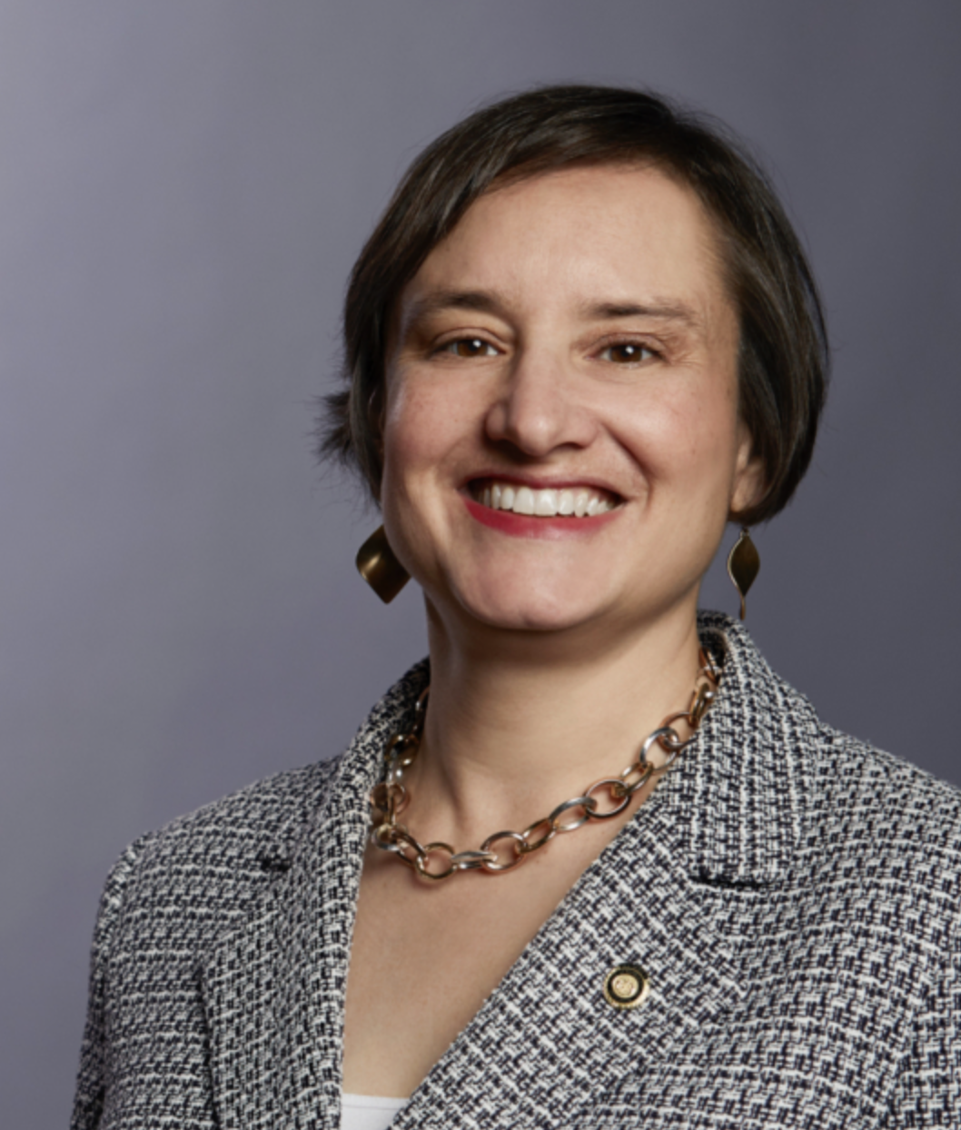 A woman with short brown hair, wearing a tweed blazer, gold chain necklace, matching earrings, and a lapel pin, smiling against a plain grey background.