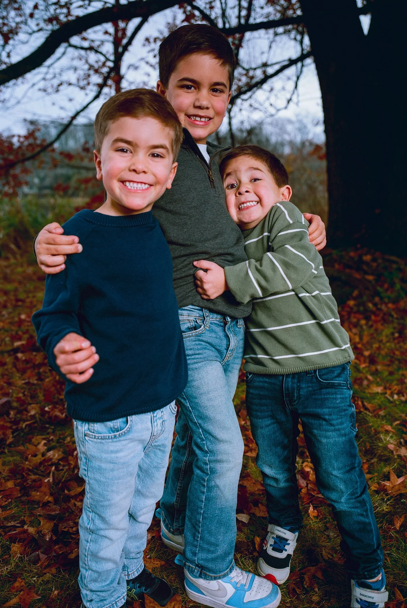 Three boys hugging outdoors in autumn, trees with fall foliage in background.