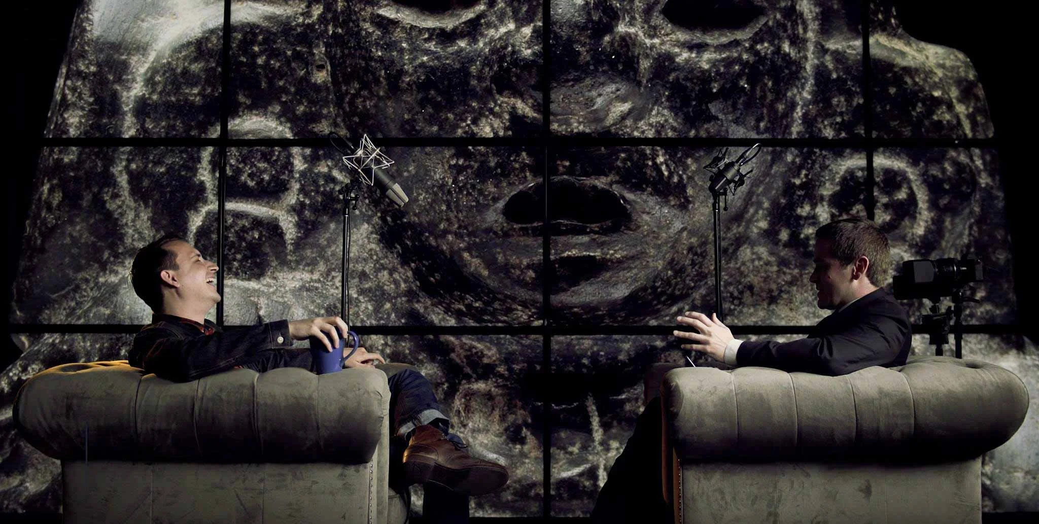 Two men are sitting on beige couches facing each other, laughing. Behind them is a large digital screen displaying a close-up image of a face, with a textured, fur-like appearance. Microphones and camera equipment are positioned near each man.