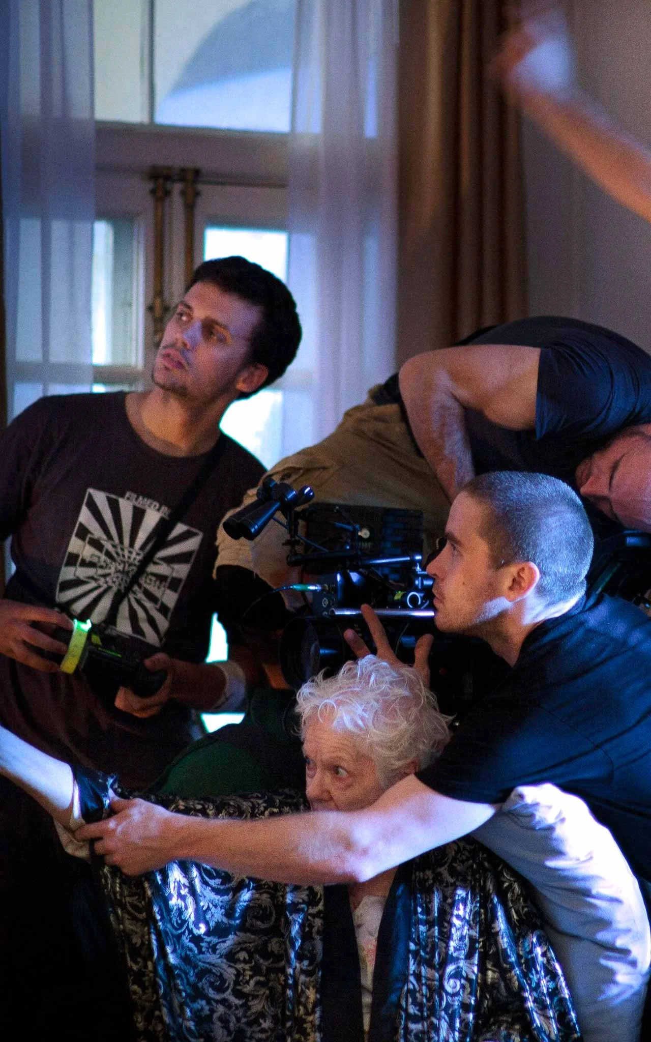A film crew is filming an elderly woman inside a room with large windows and curtains. The crew members are focused on the woman and adjusting the camera.