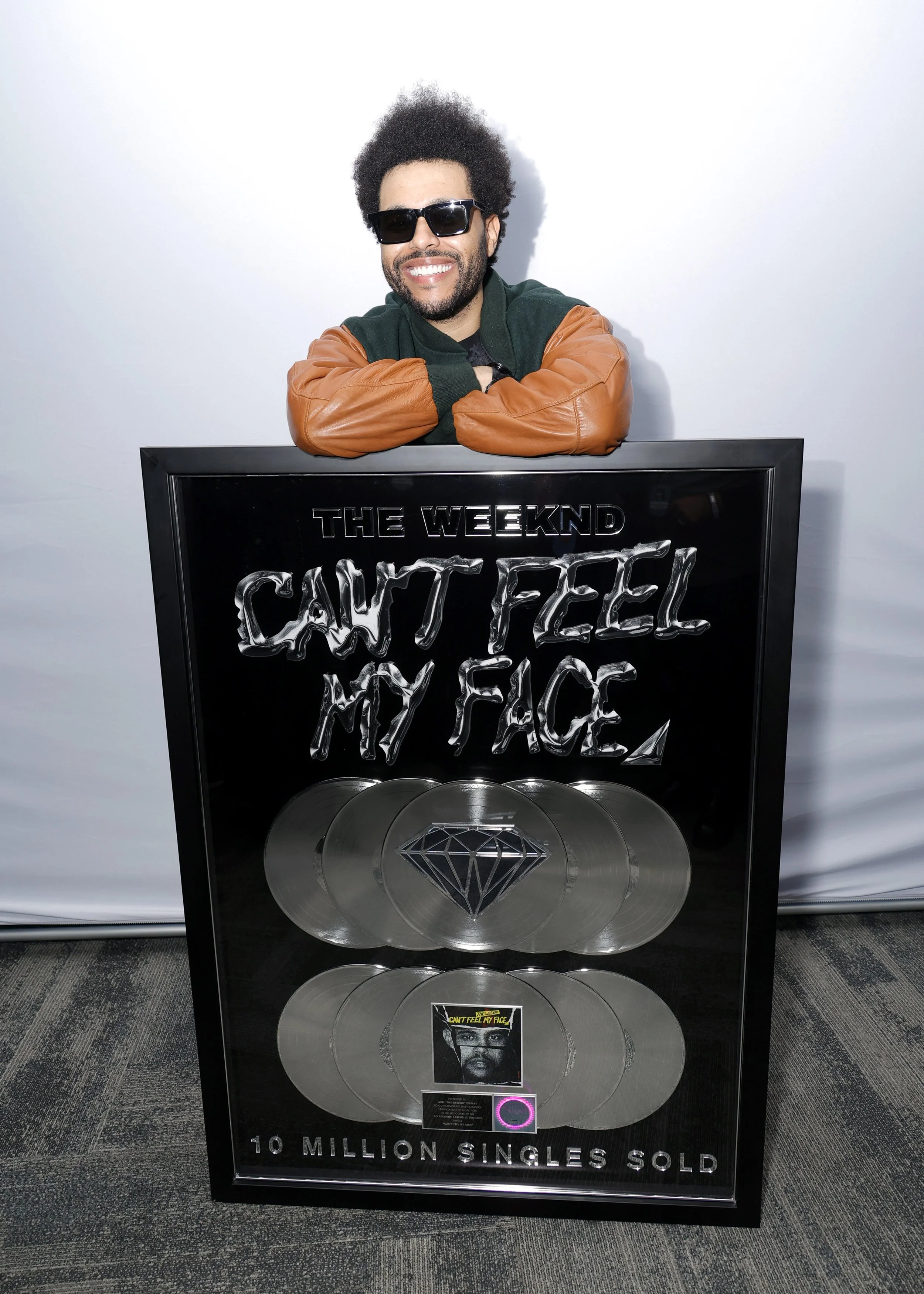 A man with sunglasses and curly hair smiling, leaning on a large black framed plaque that commemorates the album "Can't Feel My Face" by The Weeknd, with the text "10 MILLION SINGLES SOLD" at the bottom.