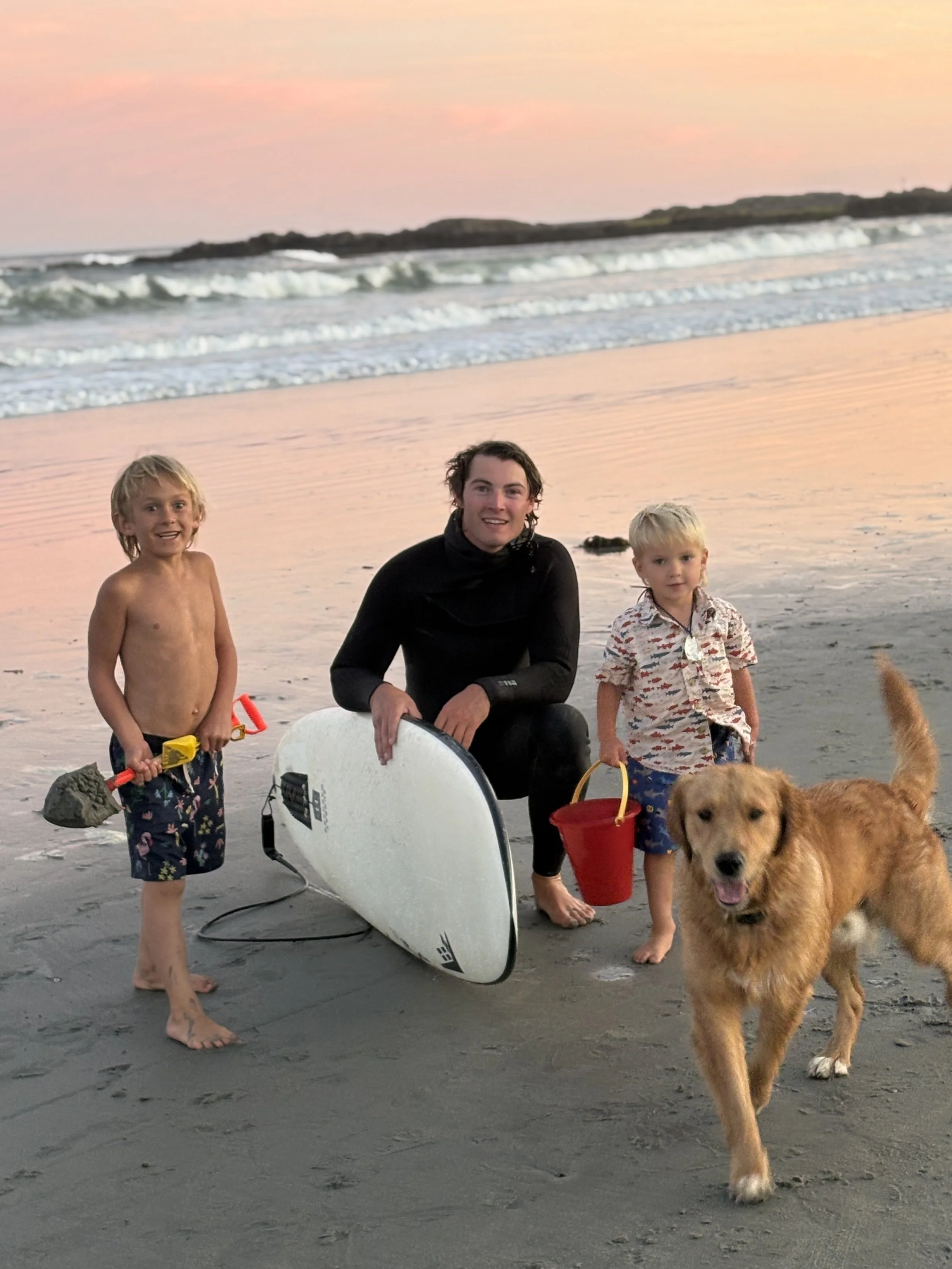A family with two young children and a dog on the beach at sunset. One child is holding a bucket and shovel, and the other is next to an adult holding a surfboard.