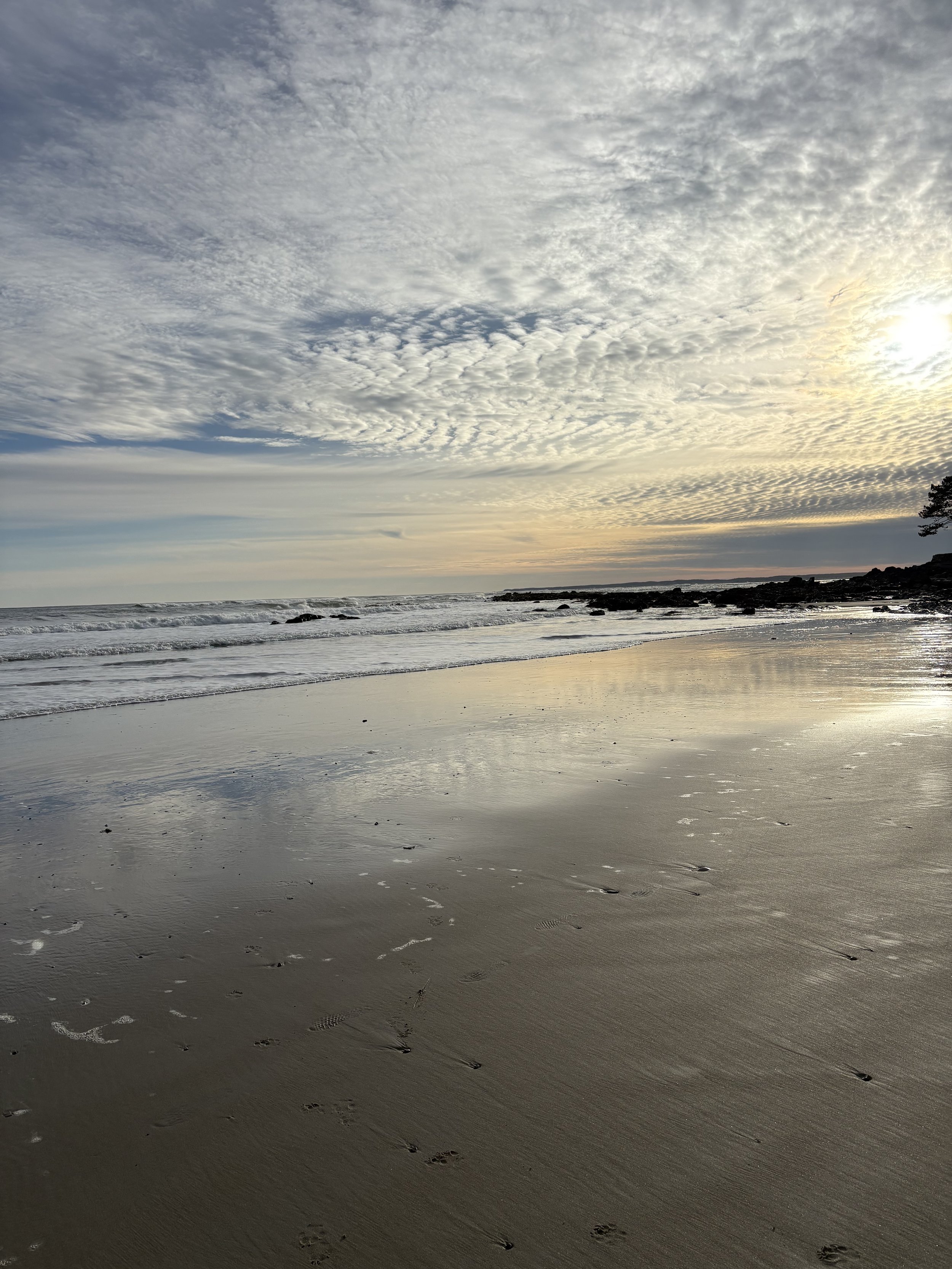 Sunset over a quiet beach with wet sand in the foreground and footprints. Gentle waves and rocks on the shoreline. Cloudy sky with sunlight.