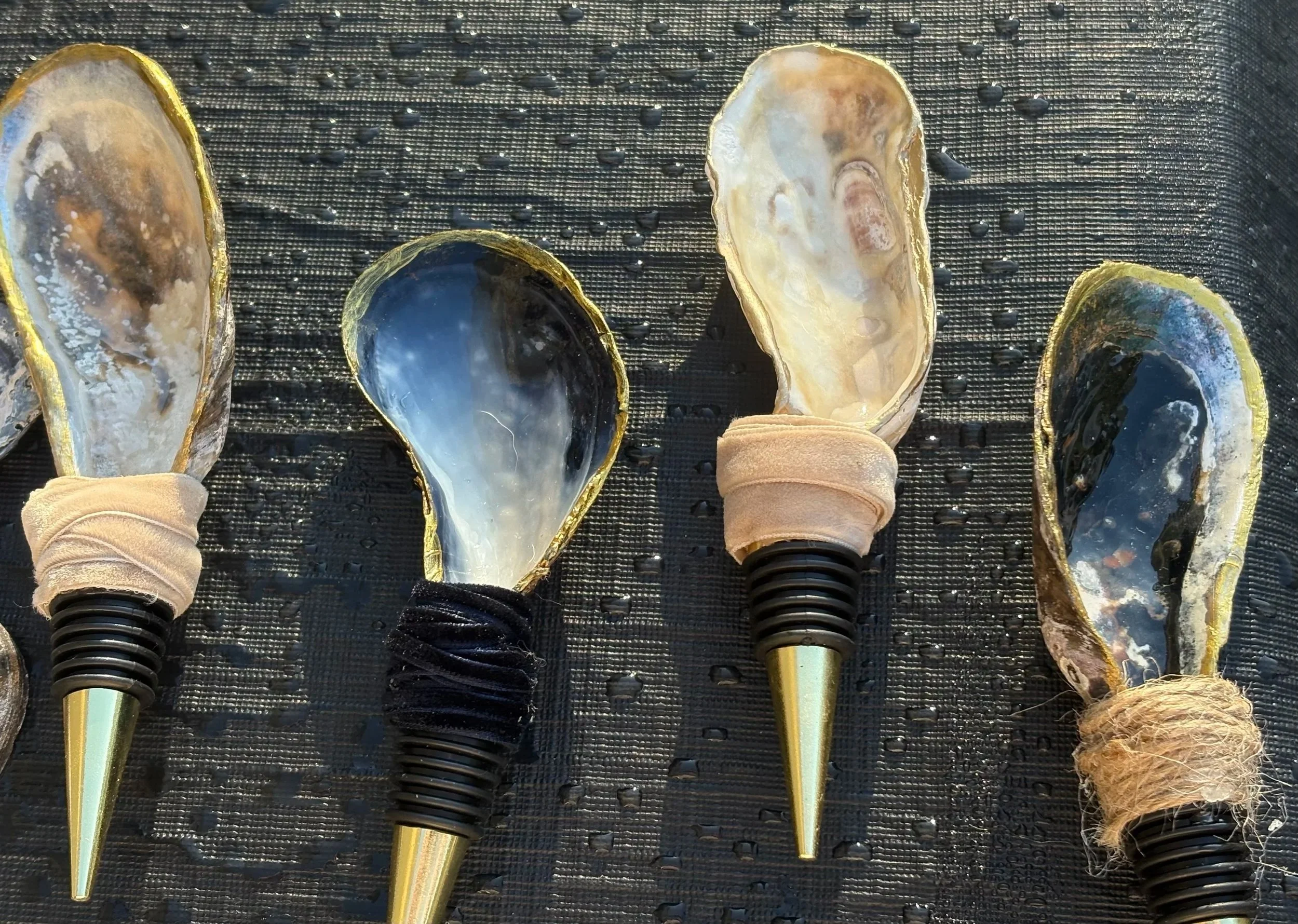 A row of four oyster shells with pointed golden tips, equipped with wine corks to serve as handles, resting on a wet, black textured surface with water droplets.