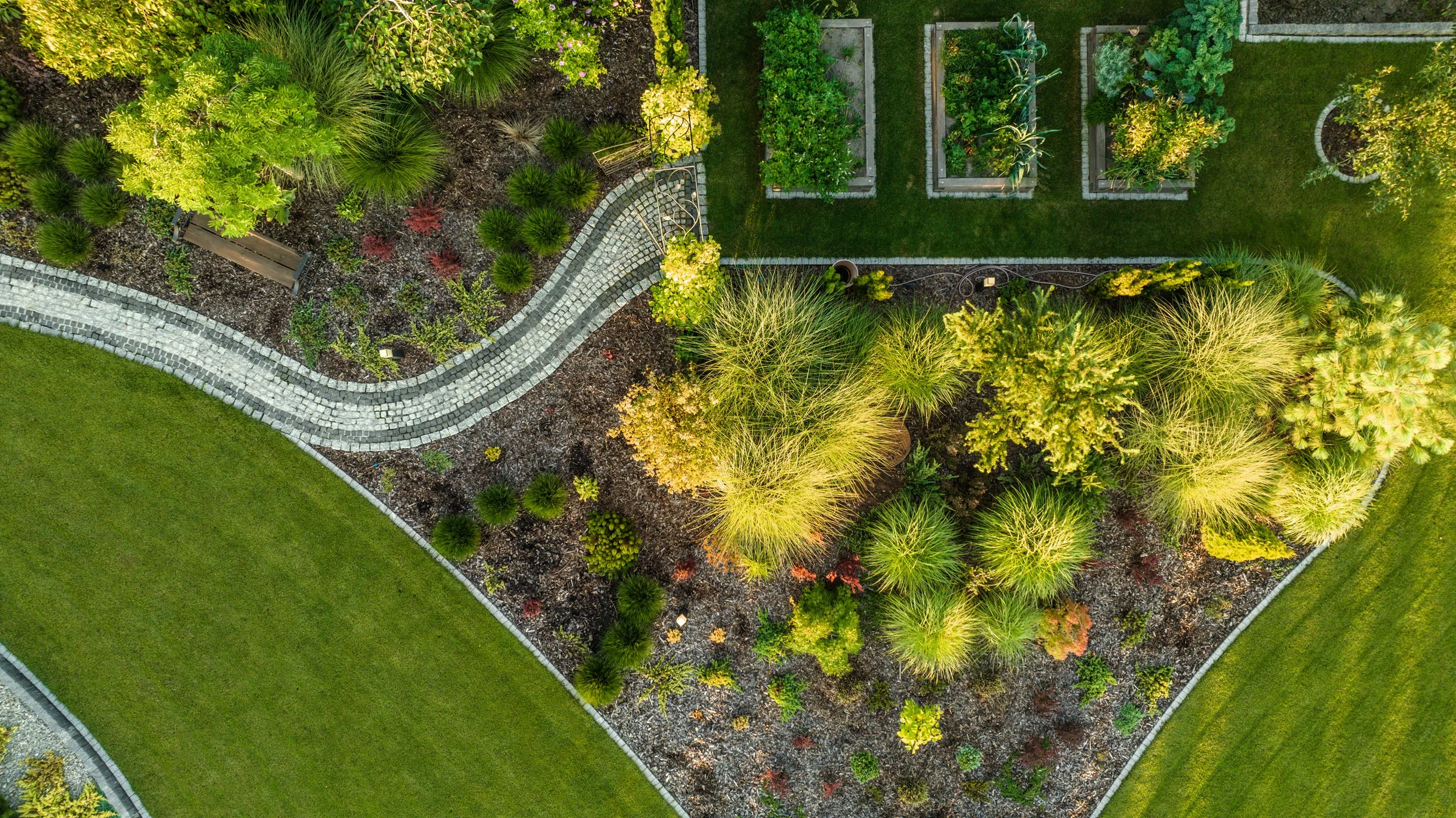An aerial view of a landscaped garden with a curved stone pathway, green grass lawns, and various plants and trees in separate garden beds.
