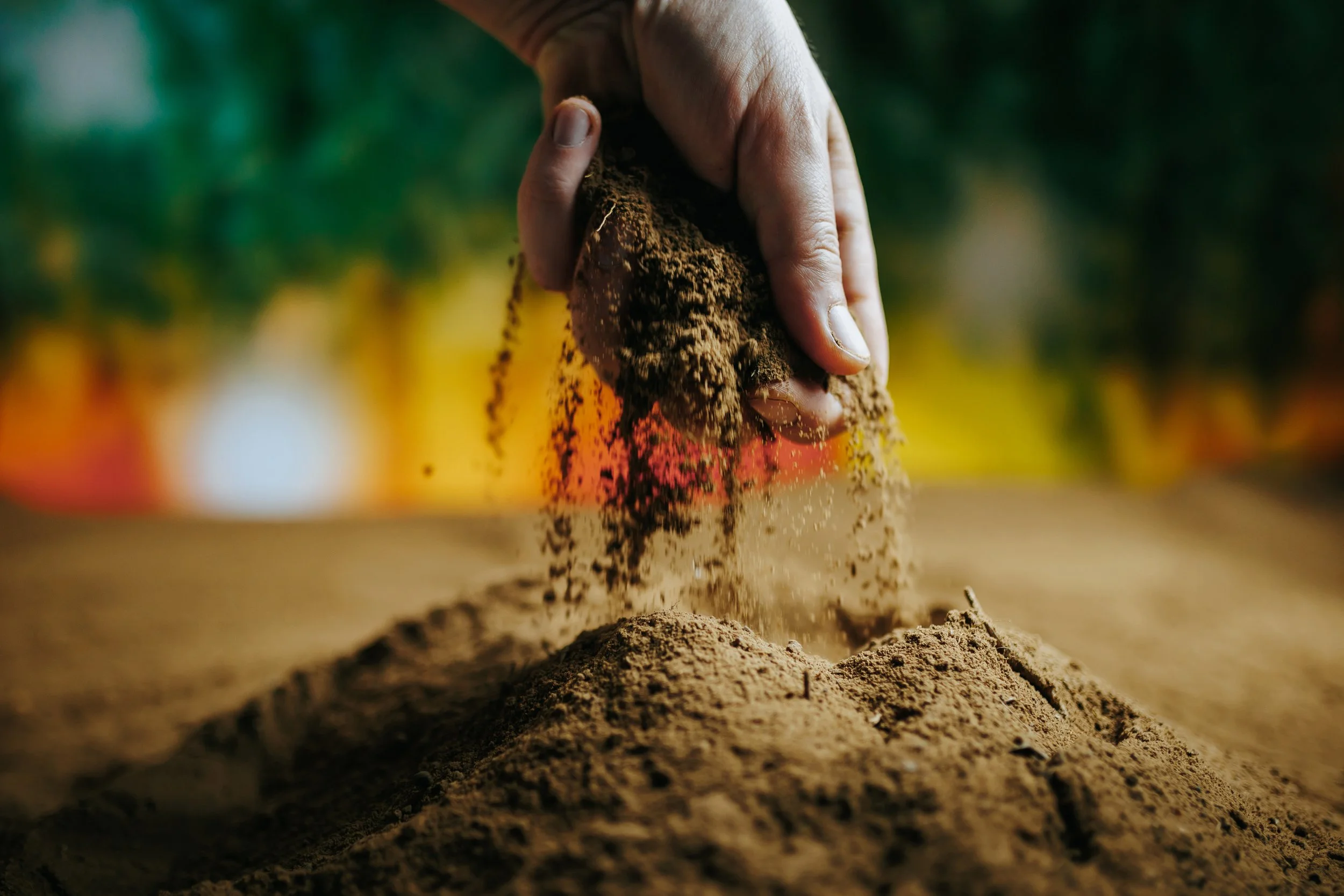 A hand sifting soil or sand, scattering it onto a surface in an outdoor setting with a blurred, colorful background.