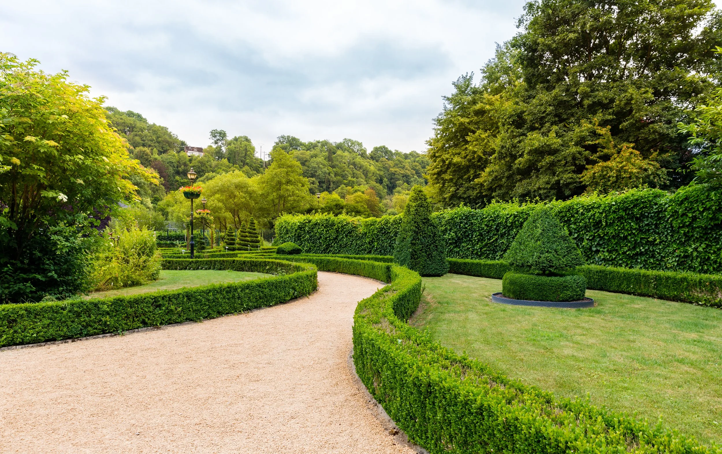 A landscaped garden with a winding gravel pathway, neatly trimmed bushes, potted conical topiary, tall trees, and a lush green hedge wall, with a backdrop of hills and a cloudy sky.