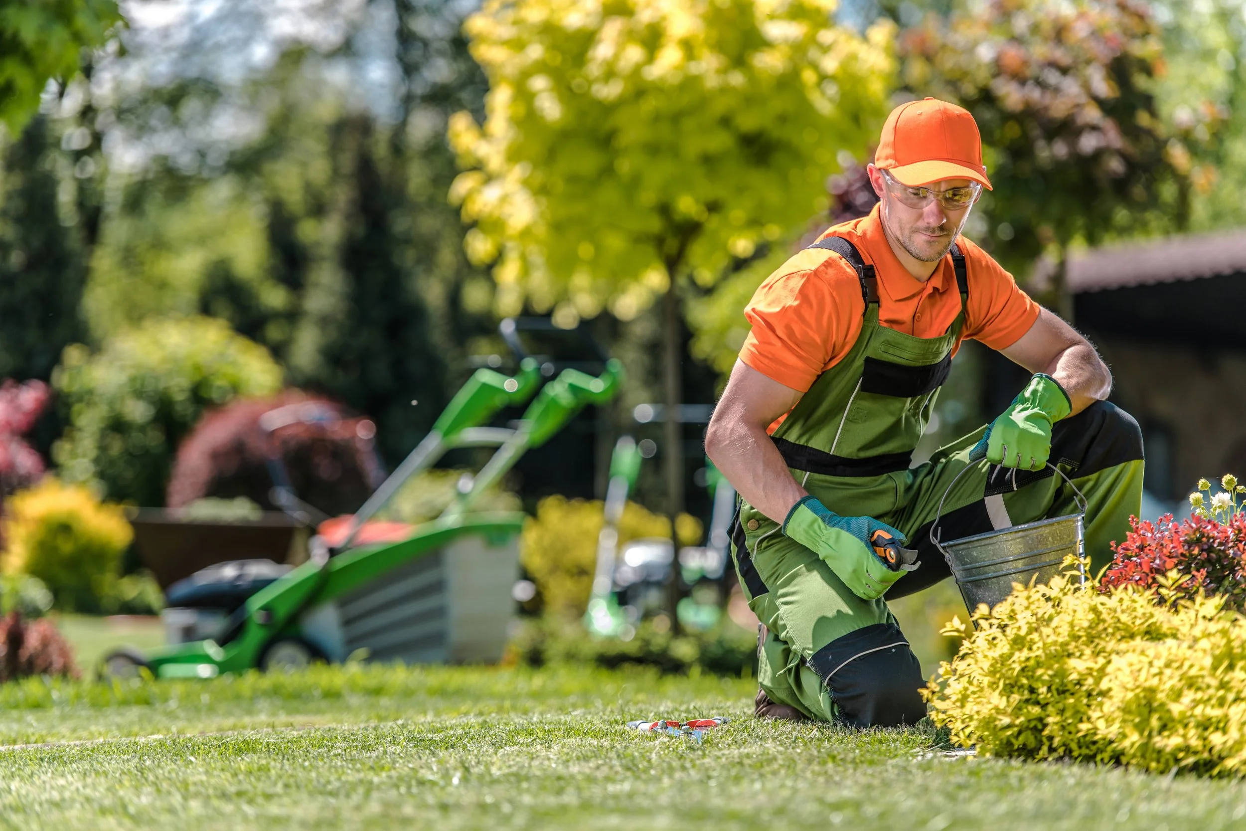 A man wearing orange and green gardening clothes, gloves, and sunglasses kneeling on the grass and tending to colorful bushes in a garden.
