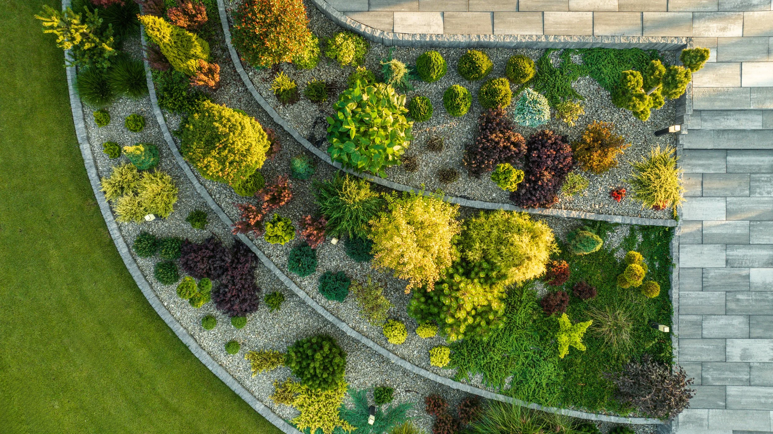 An aerial view of a landscaped garden with a variety of trees, shrubs, and plants separated by gravel paths and edged with stone borders, adjacent to a concrete patio or walkway.