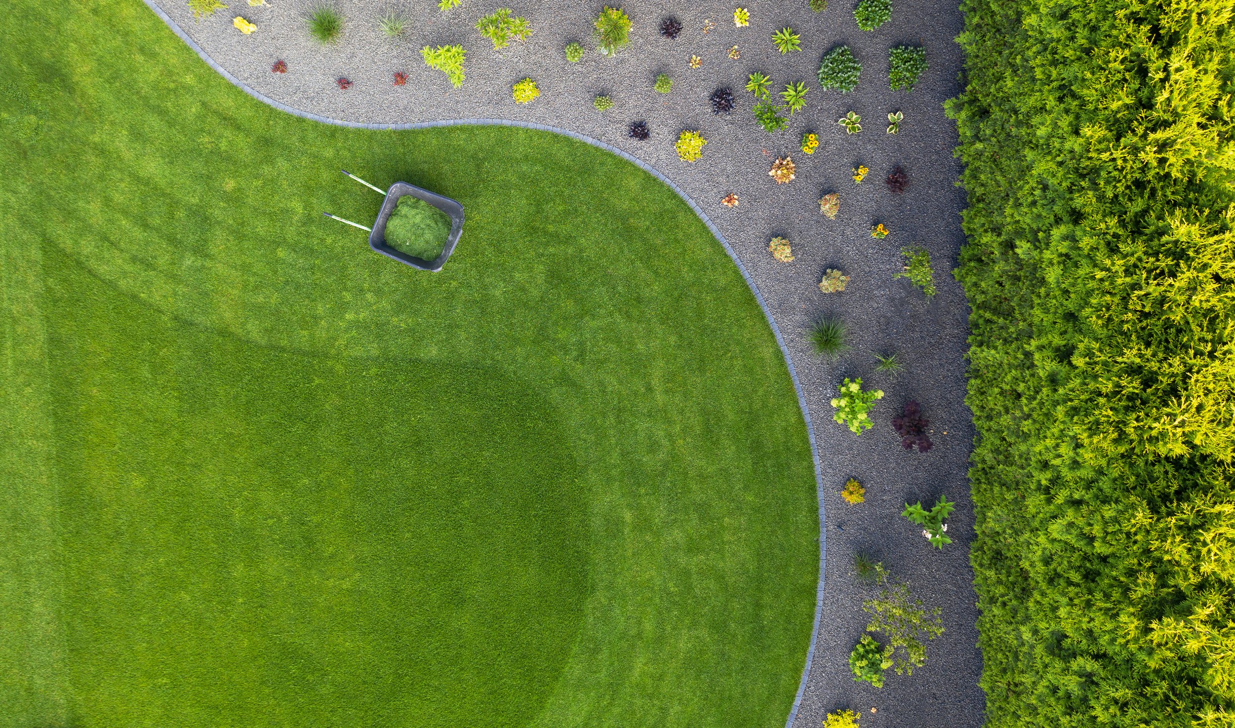 An aerial view of a neatly maintained lawn surrounded by a gravel pathway with small plants and shrubs along the edge.
