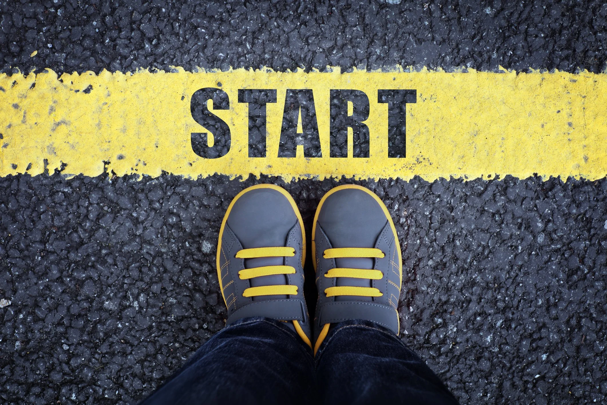 Top-down view of a person's feet in gray and yellow sneakers standing on black asphalt with a yellow line marked 'START'.