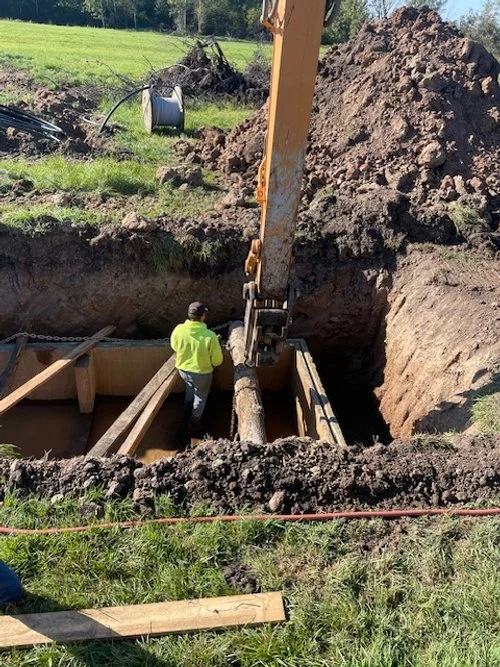 Construction worker in a high-visibility jacket working with an excavator to set a large pipe in a trench during a construction project.