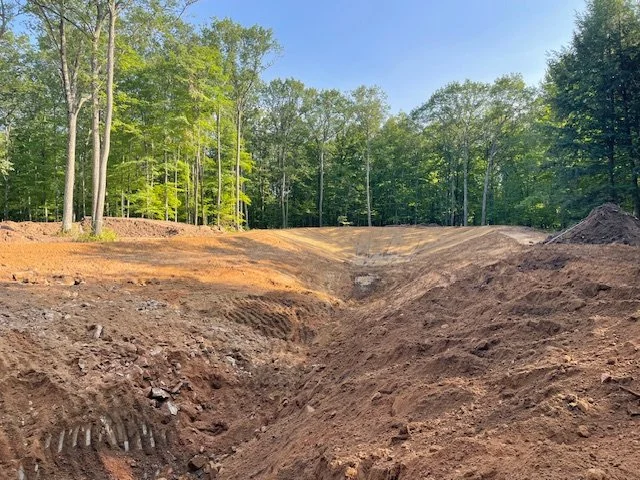 A cleared construction site with excavated dirt and a wooded area in the background under a clear blue sky.