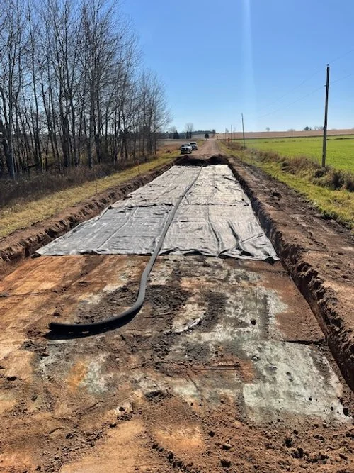 A rural dirt road under construction, with a large pipe running across it. There is a tarp or plastic cover over part of the road, and dirt trenches on both sides. A vehicle is parked in the distance, and open fields and power lines are visible under