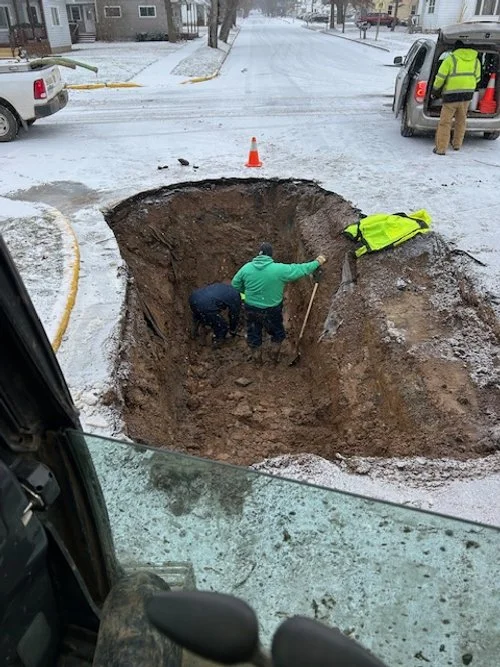 Workers are excavating a large hole in the snow-covered street, with one man inside the hole and another helping from outside. A yellow safety vest is draped over the edge, and an orange cone marks the area. Vehicles are parked nearby, and the scene 