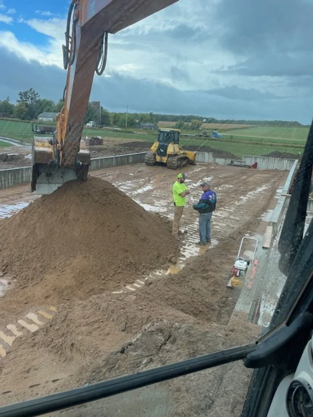 Construction site with two workers and heavy machinery, including an excavator pouring dirt and a bulldozer in the background.