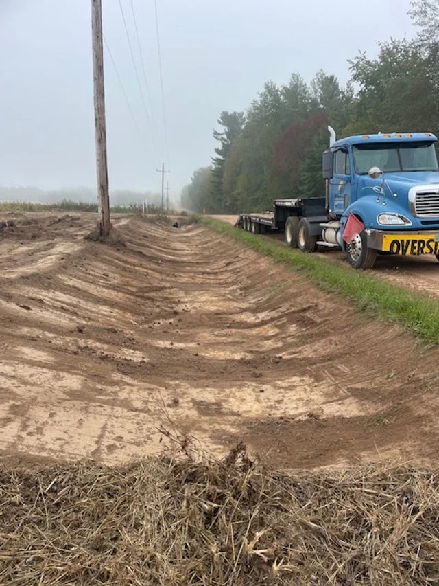 A blue semi-truck with an 'OVERSIZE' sign on the front is parked on a grassy shoulder next to dirt and construction work near a rural road with utility poles and trees in the distance.