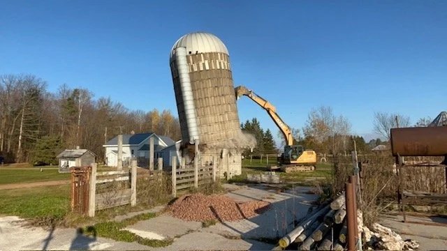 A construction site with a large damaged barn silo, an excavator, and some surrounding buildings and trees.