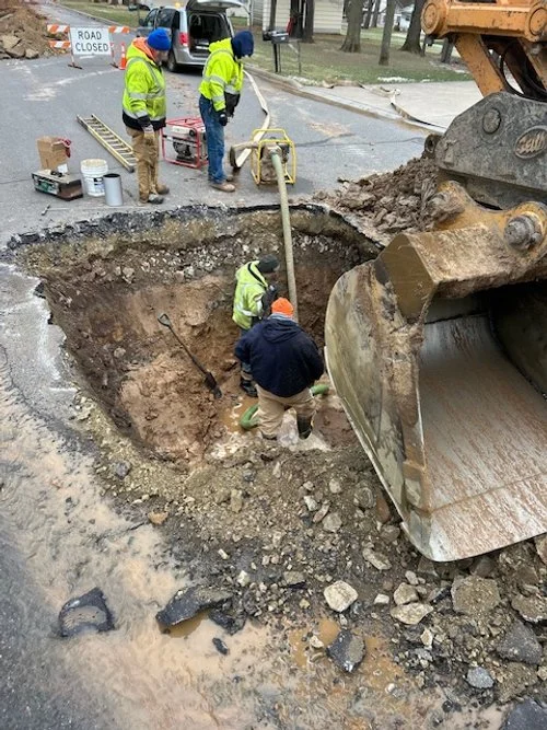Construction workers excavate a large hole on a road, with an excavator and construction tools nearby, while a 'Road Closed' sign is visible in the background.