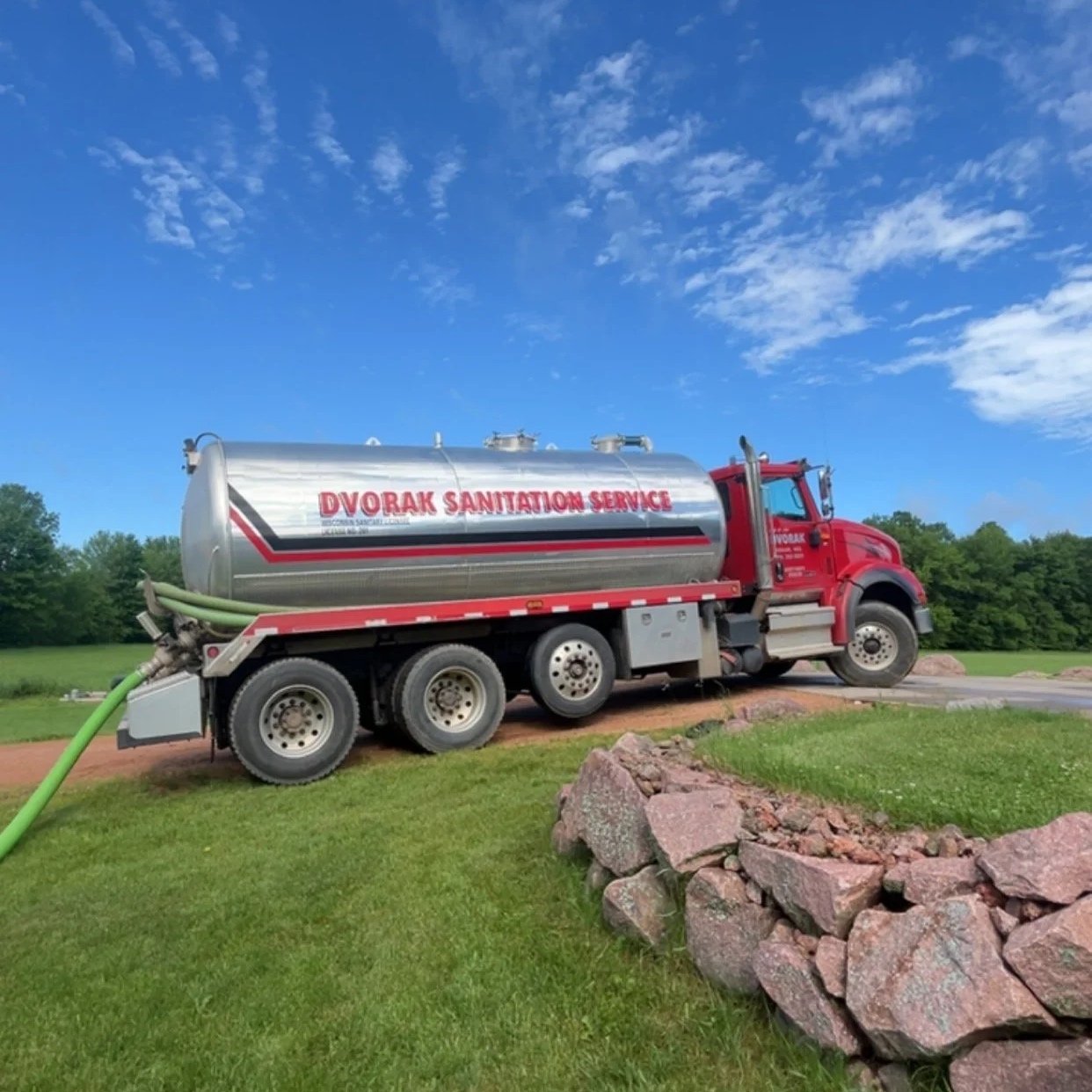 A red and silver sanitation service truck with a large tank labeled 'Dvorak Sanitation Service' parked on grass with a gravel driveway nearby and a backdrop of green trees and a blue sky with white clouds.