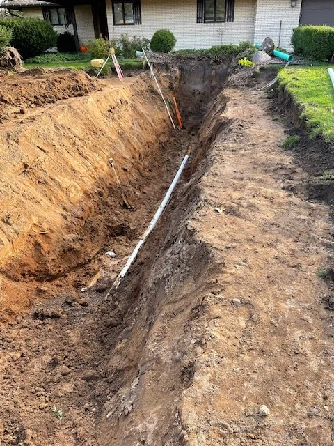 A large trench dug into the ground with measuring tools and digging equipment nearby, indicating a construction or landscaping project.