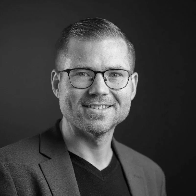 Black and white portrait of a smiling man wearing glasses, a dark blazer, and a black shirt, against a dark background.