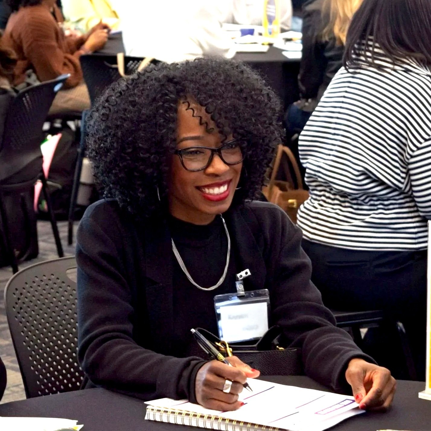 A woman with curly black hair, glasses, and red lipstick sitting at a table, smiling while writing on a notepad during a conference or seminar.