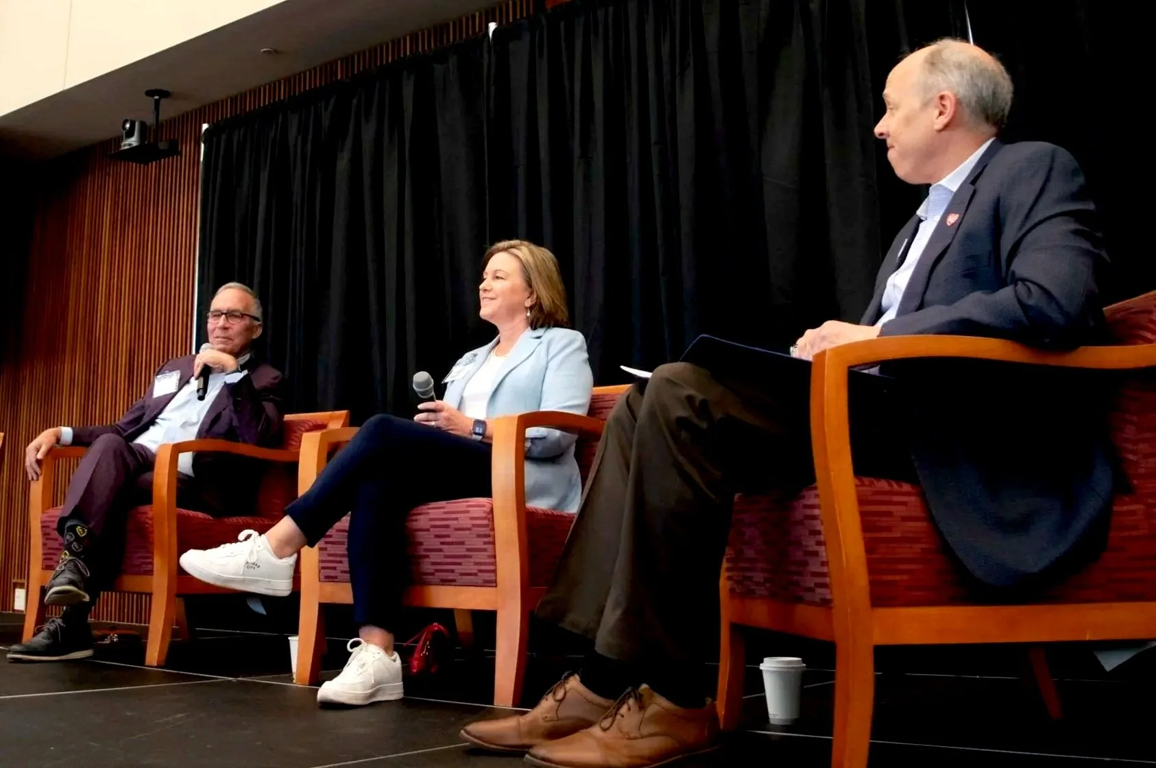 Three people sitting on stage in chairs during a panel discussion, with a black curtain backdrop.