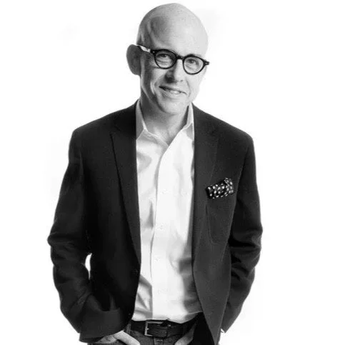 Black and white photo of a bald man wearing glasses, a dark suit jacket, and a white shirt, smiling with hands in his pockets.