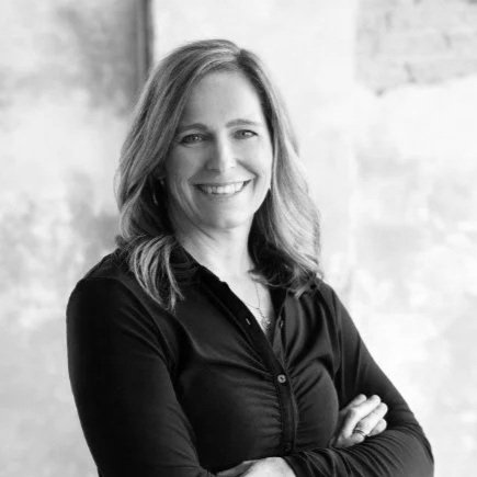 Black and white portrait of a woman with shoulder-length hair, smiling, wearing a dark button-up shirt, standing with arms crossed in front of a textured wall.