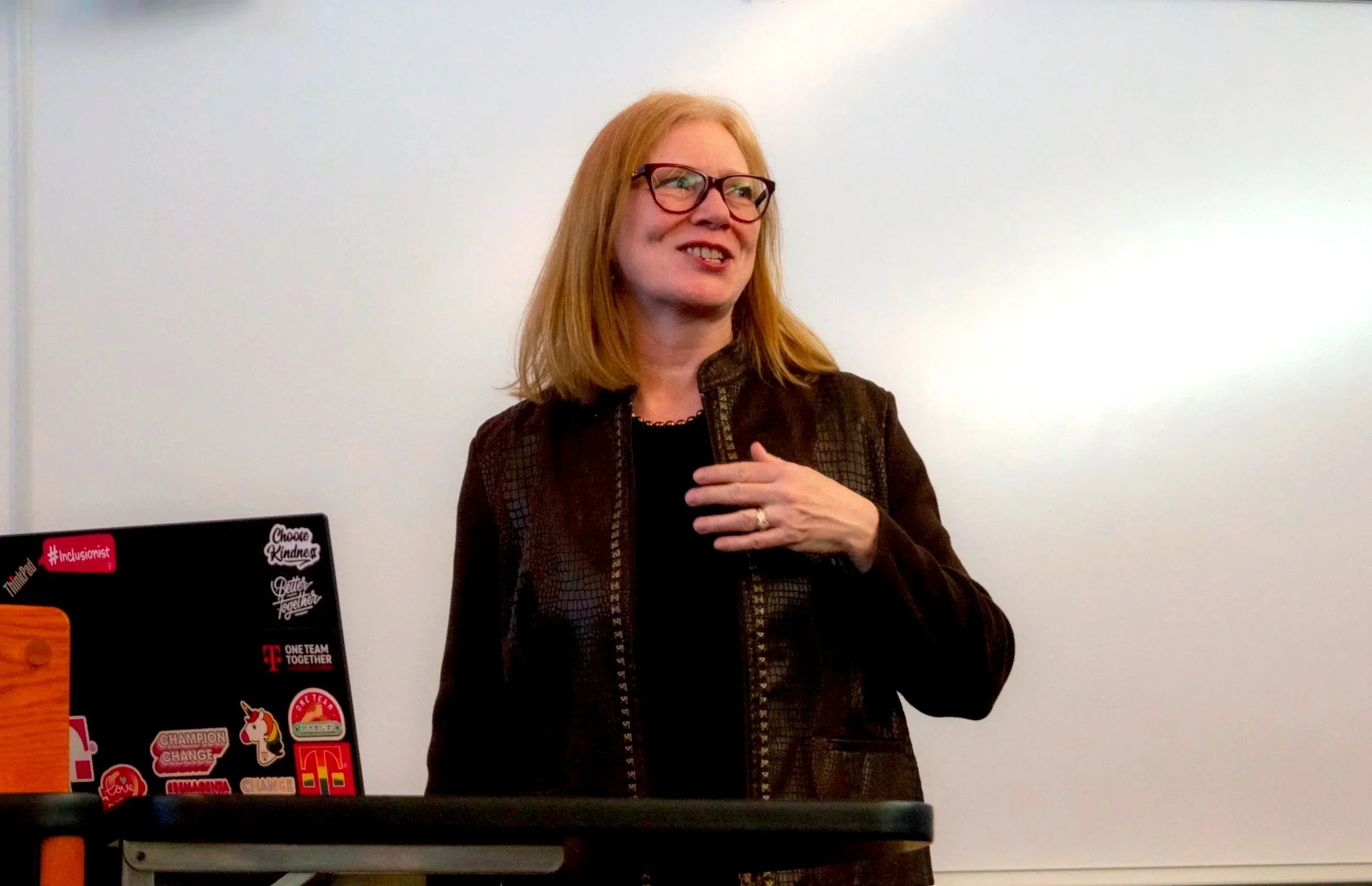 A woman with red hair and glasses standing in front of a whiteboard, wearing a brown jacket, smiling, and placing her right hand on her chest.