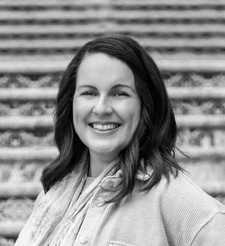 Portrait of a smiling woman with dark hair, wearing a coat and scarf, in front of steps.