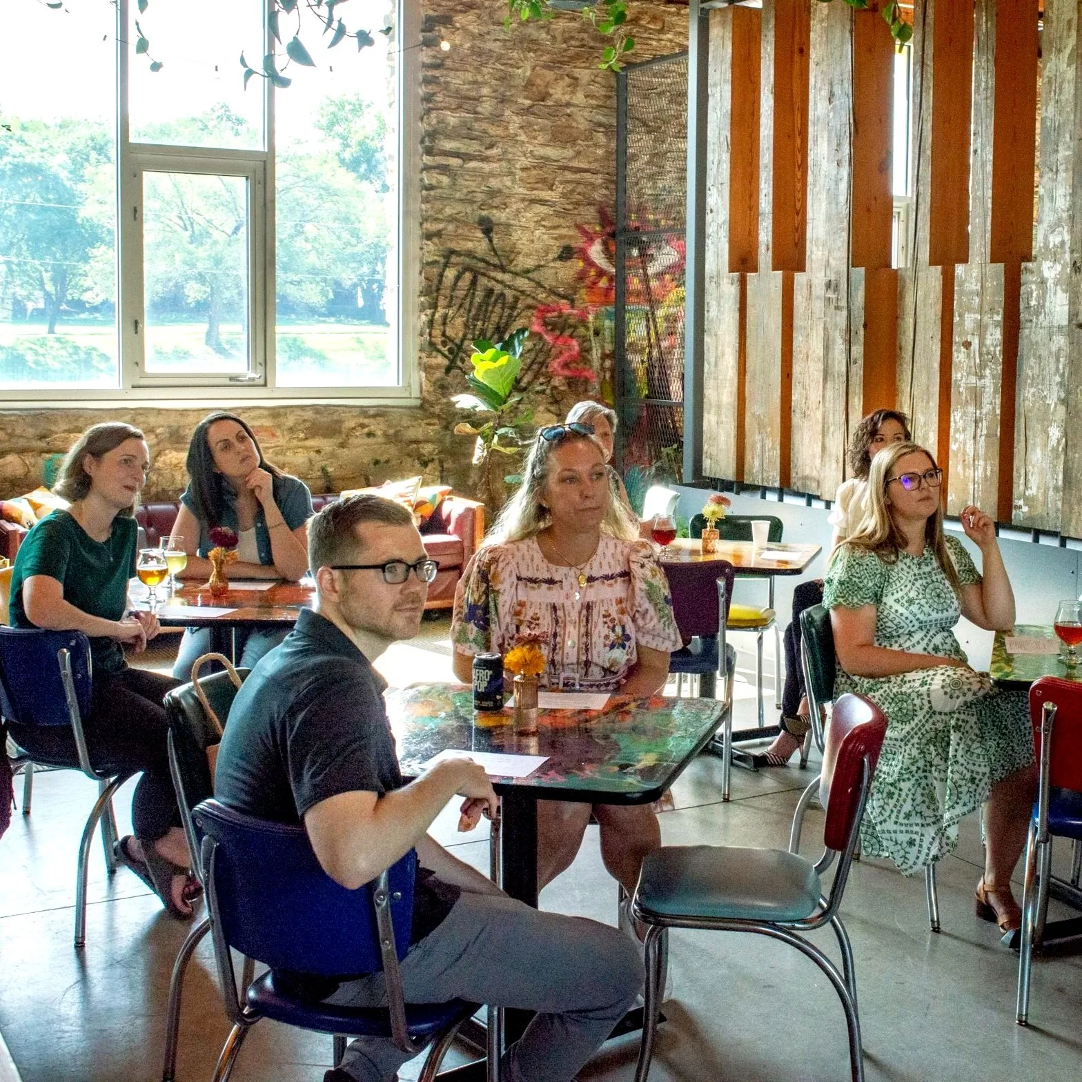 A group of people sitting in a cafe or restaurant, attentively watching something off-camera. The setting features large windows, a rustic brick wall, and wooden decor.