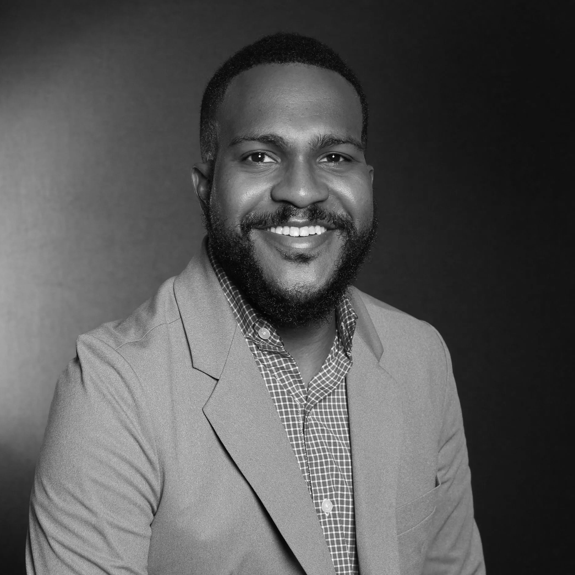Black and white portrait of a smiling man with a beard, wearing a suit jacket over a checkered shirt, against a dark background.