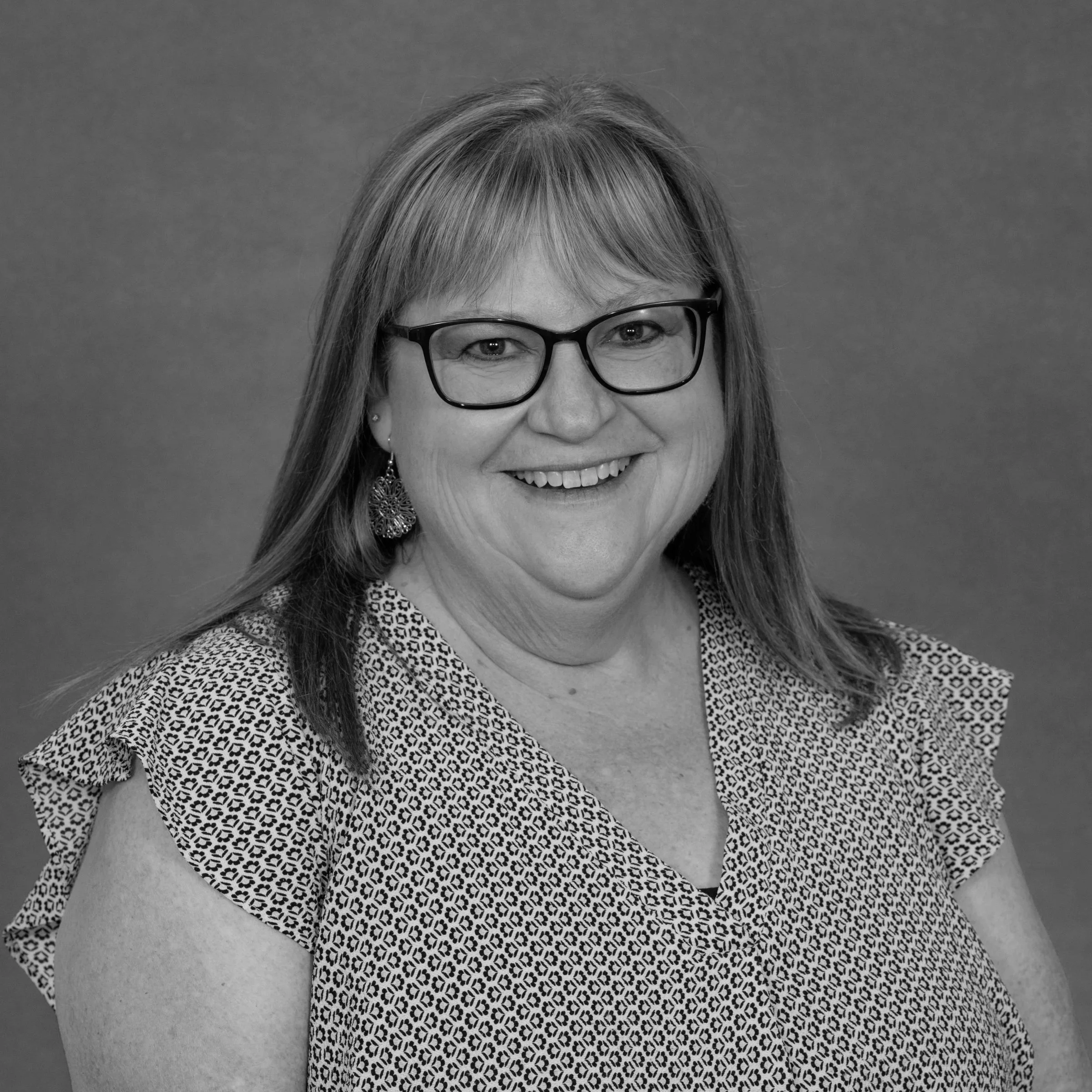 Black and white portrait of a smiling woman with glasses, earrings, and shoulder-length hair, wearing a patterned blouse, against a plain background.