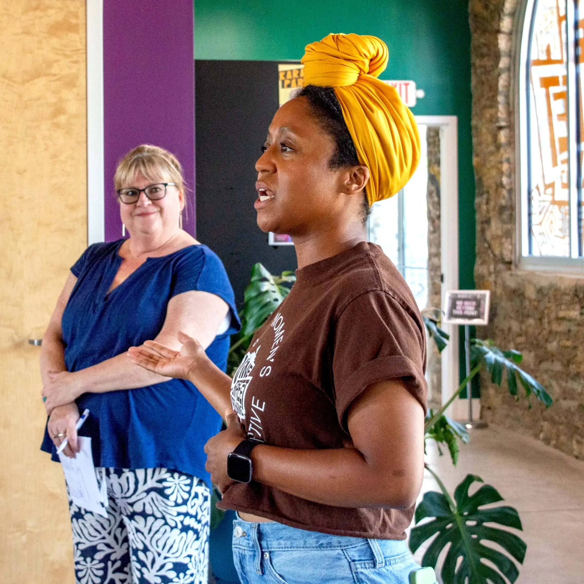 A woman with a yellow headwrap speaking to a group while another woman listens and smiles in the background.