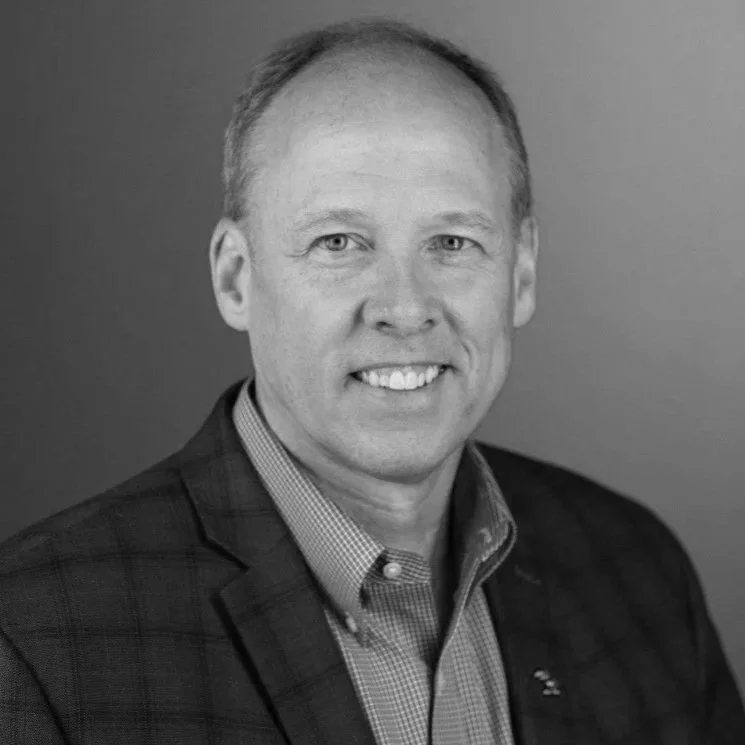A smiling man with short light-colored hair, wearing a checkered shirt and a dark blazer, posing against a plain background.