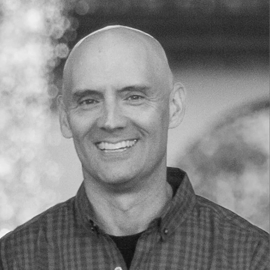 A smiling man with a shaved head, wearing a checkered shirt, standing outdoors.