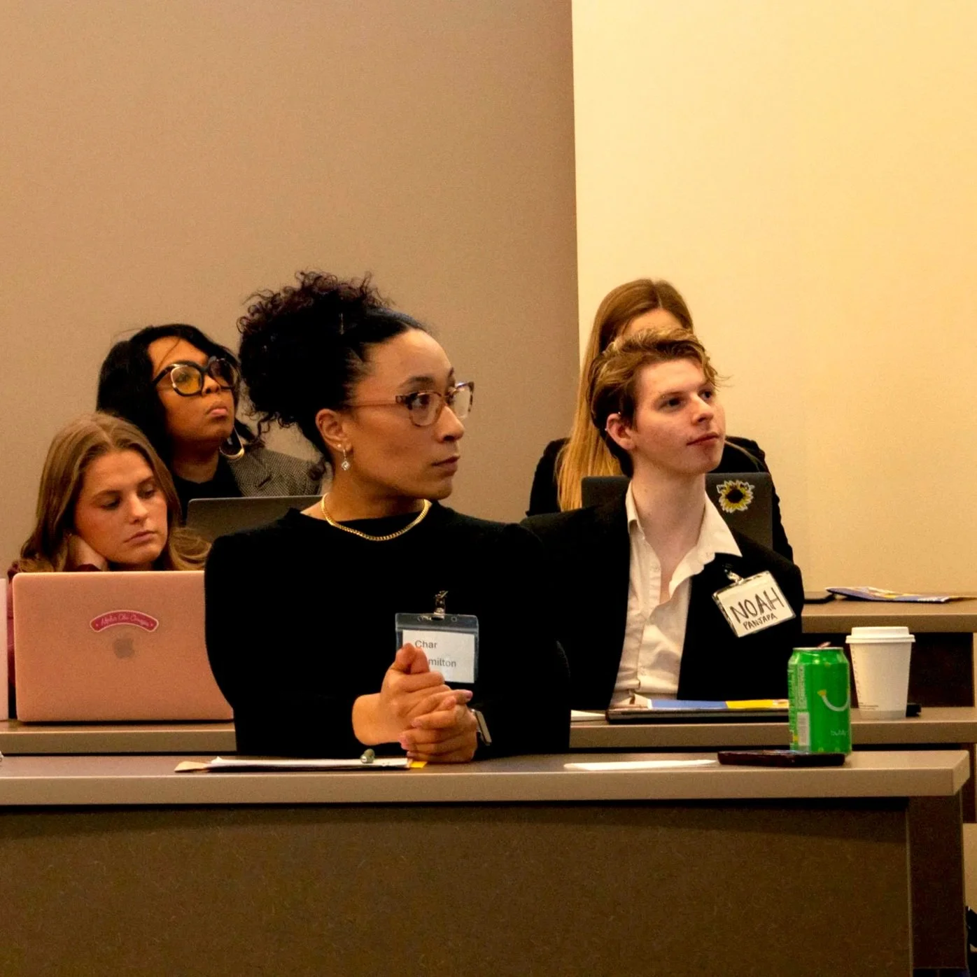 A group of diverse professionals attending a conference in a meeting room, seated at tables with laptops, drinks, and notes, listening attentively.