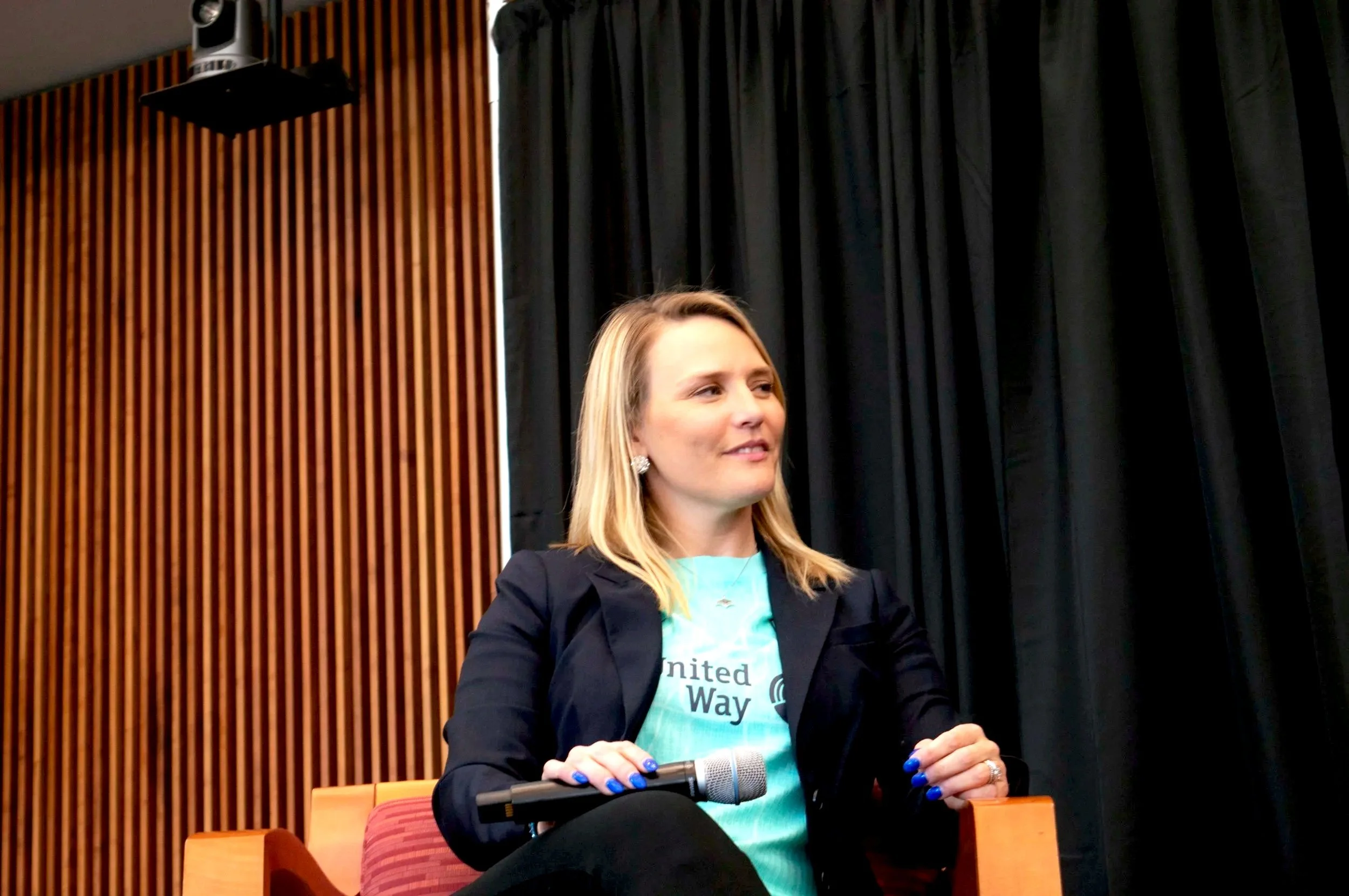 A woman with blonde hair sitting on a chair, holding a microphone, wearing a teal shirt and a dark blazer, in front of a black curtain with wooden paneling behind her.