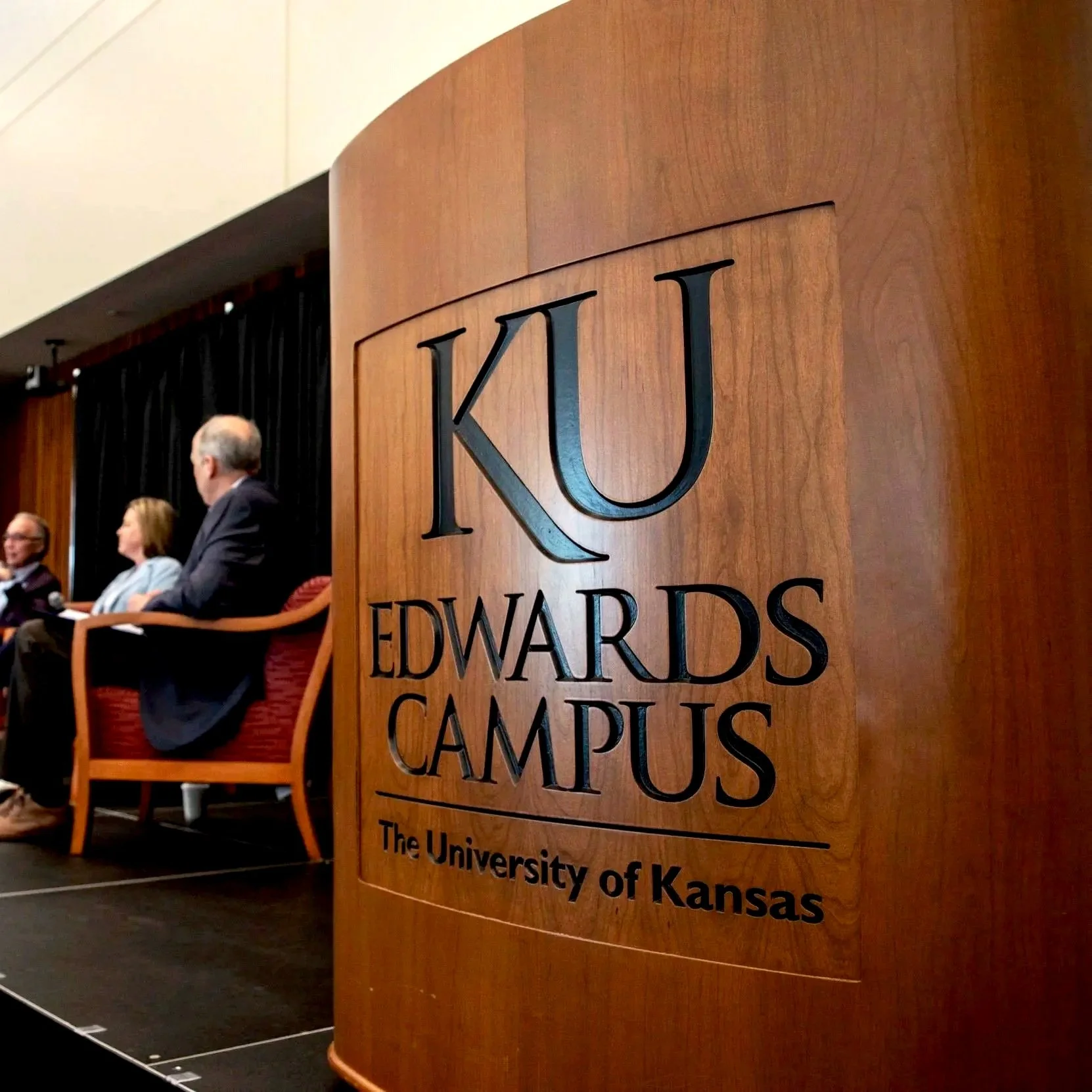 A wooden panel with the logo and name of KU Edwards Campus at the University of Kansas in the foreground. In the background, there are three people sitting on stage chairs, participating in a panel or discussion at a conference or event.
