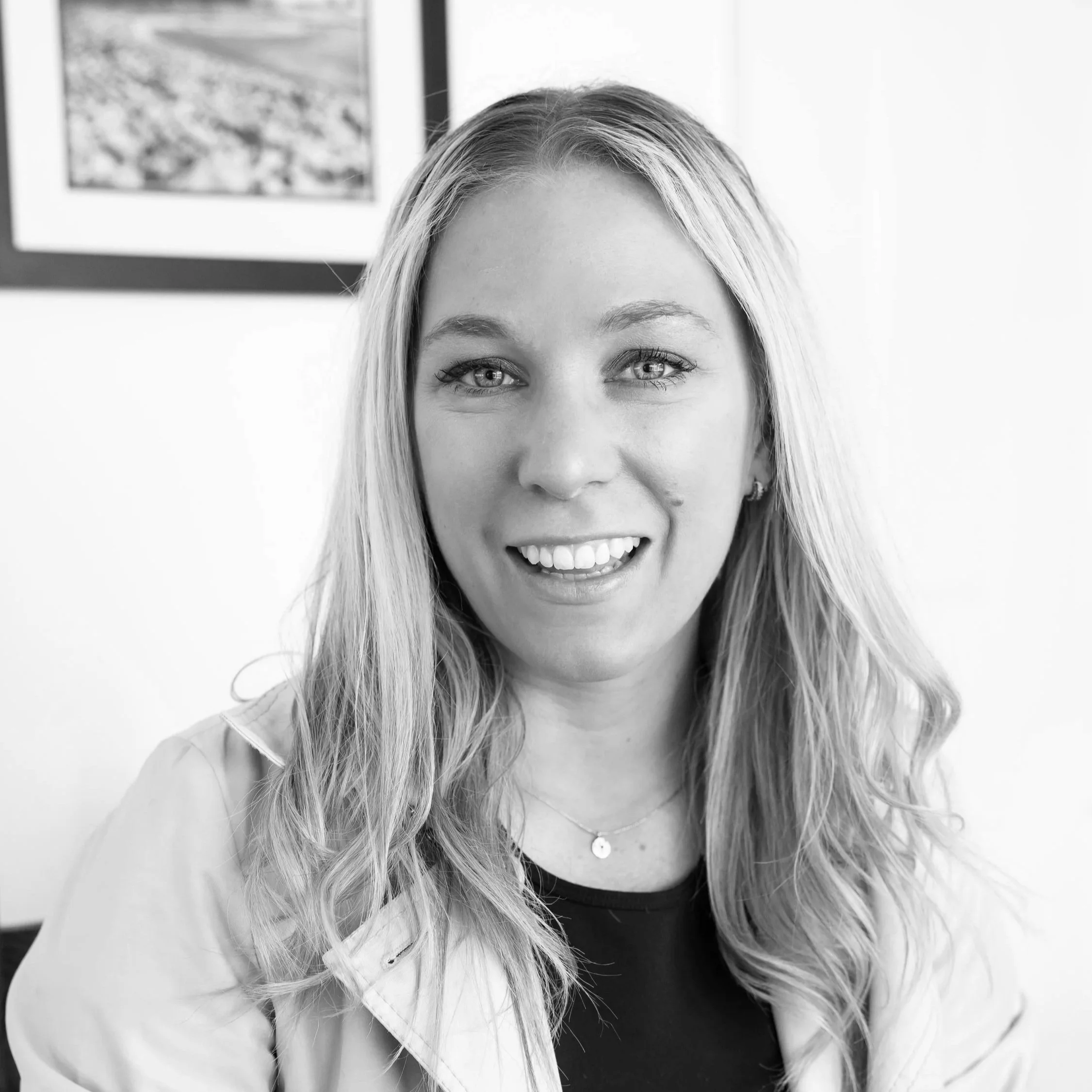 A black and white photo of a smiling woman with long wavy hair, wearing a light-colored jacket and a necklace, indoors near a wall with framed artwork in the background.