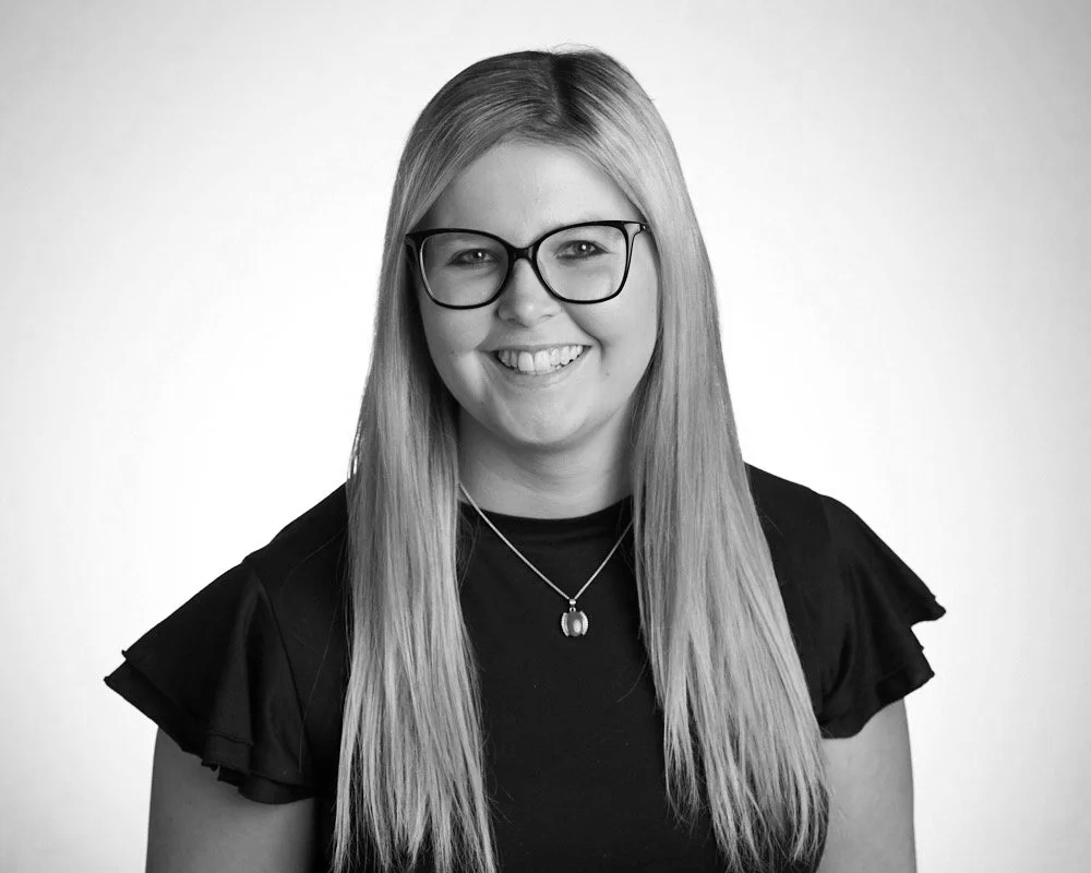 Black and white portrait of a young woman with long straight hair, wearing glasses and a necklace, smiling, against a plain background.