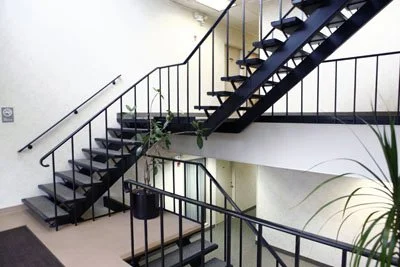 Indoor staircase with black metal railing and steps, located in a well-lit area with plants and a hallway visible.